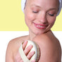 A smiling person with a towel-wrapped head gently brushes their bare shoulder with the Afterspa Massage & Detox Brush, enjoying exfoliation against a yellow and white background.