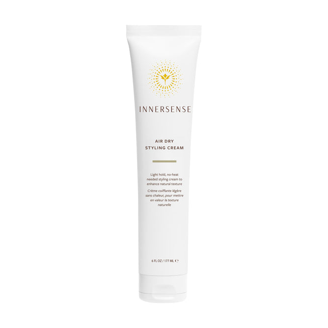 A white tube of Innersense Organic Beauty Air Dry Styling Cream stands upright, adorned with gold and black text, a yellow sunburst logo, and product information in both English and French. This lightweight smoothing cream from Innersense Organic Beauty provides frizz control for effortless, heat-free styling.