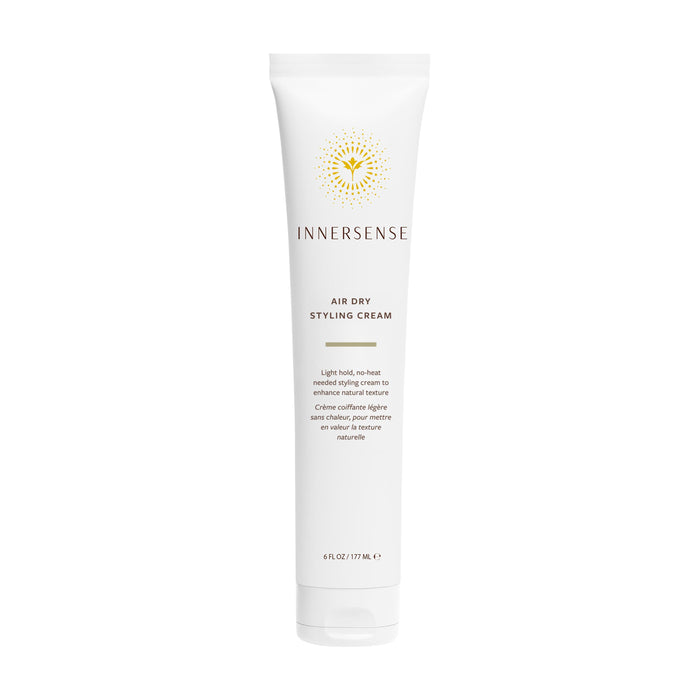 A white tube of Innersense Organic Beauty Air Dry Styling Cream stands upright, adorned with gold and black text, a yellow sunburst logo, and product information in both English and French. This lightweight smoothing cream from Innersense Organic Beauty provides frizz control for effortless, heat-free styling.