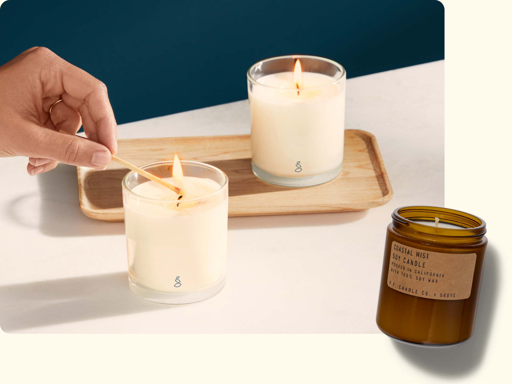 A hand lights a white candle in a glass jar with a matchstick next to another lit candle on a wooden tray. An amber jar labeled Coastal Mist Soy Candle is displayed in the bottom right corner.