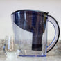 The New Wave Enviro Alkaline Pitcher Water Filter, featuring measurement markings and a black filter, sits partly filled on a kitchen counter beside a glass of water—ideal for alkalizing your water at home.