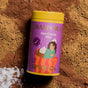 A yellow canister labeled Diaspora Spice Co. Hot Cocoa Drink Mix rests on a bed of cocoa powder. The label depicts a woman lounging in a mug of cocoa topped with whipped cream. Surrounding the canister are scattered cocoa and powdered ingredients, highlighting this hot cocoa blend made with single-origin spices and sweetened with Madhur Jaggery.