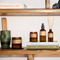 A wooden shelf displays brown glass candles, amber glass bottles, a P.F. Candle Co. Reed Diffuser for flame-free fragrance, a green ceramic holder, and books—including All-American Ads and John Cage—A Mycological Foray. A camera sits on the top shelf.