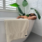 A man relaxes in a white bathtub in a sunlit bathroom with green plants nearby. An Anact Hemp and Organic Cotton Wash Cloth Bath Sheet hangs over the tub’s edge, while sunlight streams through white window shutters.