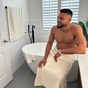 A shirtless man with a short beard sits on a white bathtub in a modern bathroom, smiling and wearing an Anact Hemp and Organic Cotton Wash Cloth Bath Sheet. A matching towel hangs on the wall, and there’s a large plant nearby.