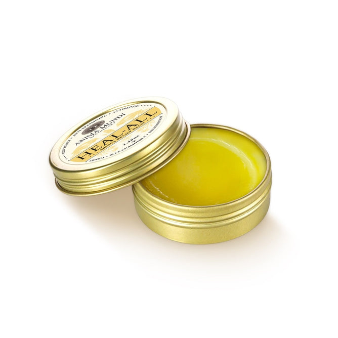 A small, round open tin of Anima Mundi HEAL-ALL Arnica Relief Balm, Nourish, Soothe + Repair with yellow balm inside and its lid partially on top, set against a white background.