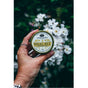 A hand holds a tin of Anima Mundi HEAL-ALL Arnica Relief Balm, Nourish, Soothe + Repair in front of blurred white flowers and green foliage, highlighting the nourishing botanical remedy tin and hand in the foreground.