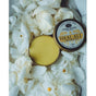 An open tin of Anima Mundi HEAL-ALL Arnica Relief Balm, Nourish, Soothe + Repair sits surrounded by white flower petals with water droplets; the lid with product details is placed beside the balm.
