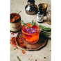 A vibrant orange-red MANGOSTEEN HIBISCUS, Beauty Tonic by Anima Mundi sits on a rustic plate, garnished with a purple edible flower and rosemary, surrounded by hibiscus powder, a dropper bottle, and cocktail tools.