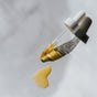 A glass dropper with a white cap, filled with yellow Little Seed Farm Antioxidant Serum - Protect + Revitalize, rests on a light gray surface. Beside it, a small spill of the serum creates a soft shadow.