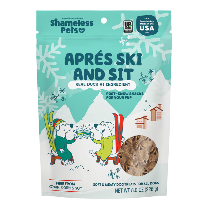 A blue and white bag of Shameless Pets Dog Treats - Après Ski and Sit, made with real duck, features two cartoon dogs in winter outfits snacking together amid a snowy mountain and tree backdrop.