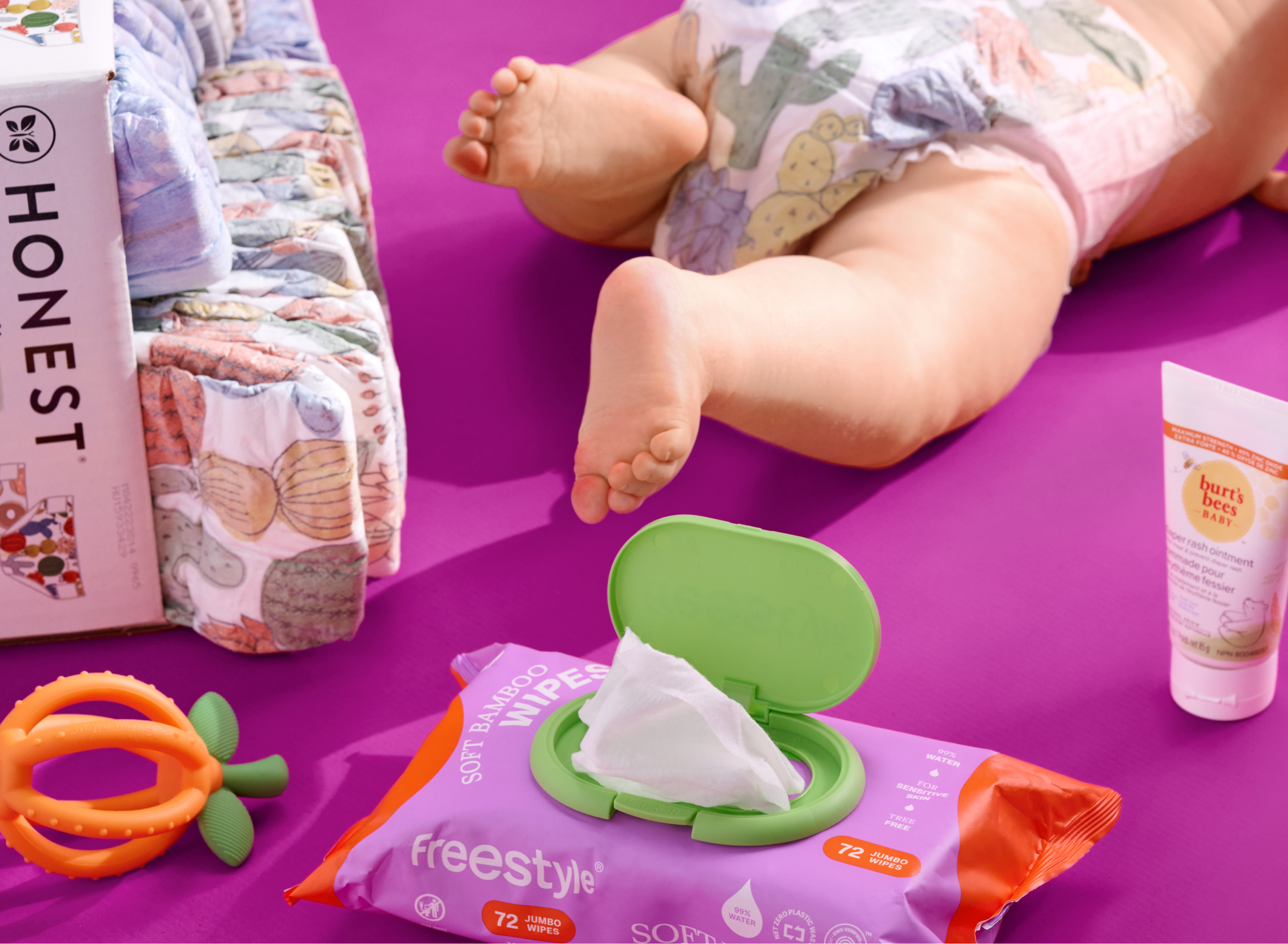 A baby lies on a purple surface wearing a diaper, surrounded by diaper packs, a pack of bamboo wipes with the lid open, a tube of cream, and an orange teething toy.