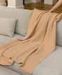 Hands draping a tan, temperature-regulating AU Baby Adult Popcorn Blanket - Henna over a light gray sofa.