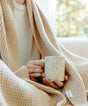 A person wrapped in a light brown, plant-dyed Adult Popcorn Blanket - Henna by AU Baby, holding a speckled mug.
