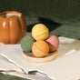 Four vibrant bath bombs from the Pacha Soap Co. Bath Bomb Set - Autumn Heirlooms Collection are neatly stacked on a small dish atop open notebooks. In the background, a pumpkin-shaped mug filled with a warm beverage adds to the cozy atmosphere, all arranged on a soft green surface for the perfect autumn-inspired scene.