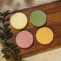 Four round, handcrafted shower steamers from the Autumn Heirlooms Collection by Pacha Soap Co., in shades of yellow, green, pink, and orange—each featuring a delicate leaf design—are arranged on a wooden board with green leaves beside them, set atop a textured beige cloth.