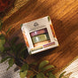 A box of Pacha Soap Co. Shower Steamer Set - Autumn Heirlooms Collection rests on a wooden surface with green leafy branches and a woven mat nearby. The packaging features four vibrant steamers in autumn-inspired scents, each designed to bring a delightful autumn fragrance to your shower experience.