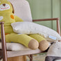 A large yellow knitted dog plush sits on a beige armchair, holding an Avocado Toddler Pillow. The background shows a green wall, with part of another pillow visible in the corner.