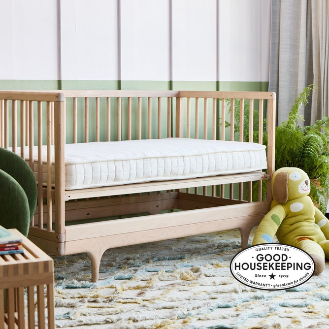 An Avocado Green Mattress Eco Organic Crib Mattress with a white cover is placed inside a wooden baby crib in a cozy nursery. Nearby, there is a plush green doll and some potted plants. A Good Housekeeping quality seal appears in the bottom right corner.