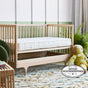 An Avocado Green Mattress Eco Organic Crib Mattress with a white cover is placed inside a wooden baby crib in a cozy nursery. Nearby, there is a plush green doll and some potted plants. A Good Housekeeping quality seal appears in the bottom right corner.