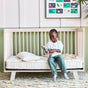A young child sits on a daybed, smiling while putting on gray socks. The bed features an Eco Organic Crib Mattress by Avocado Green Mattress and pillows, with colorful animal art hanging on the wall behind.