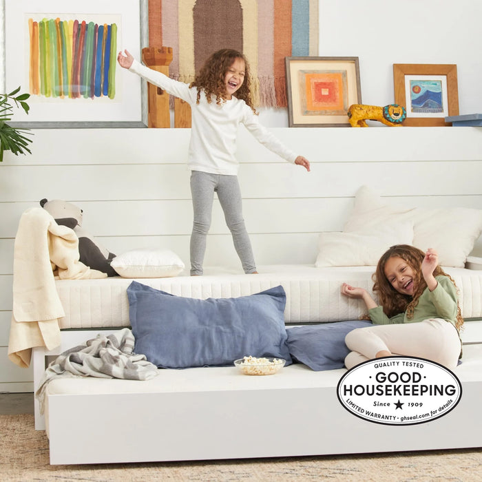 Two young girls smile and play in a bright, cozy bedroom with white walls, blue pillows, and colorful framed art. One stands on a bed topped with the Avocado Green Mattress Eco Organic Kids Mattress, while the other lounges comfortably on a trundle bed. A Good Housekeeping seal appears in the corner.