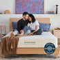 A smiling couple sits on an Avocado Green Mattress Eco Organic Mattress, leaning close together in a cozy, modern bedroom. A Good Housekeeping Bedding Awards badge is displayed in the bottom right corner.