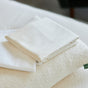 A neatly folded Avocado Organic Indian Cotton 400 TC Pillow Case Set rests on a textured cream pillow with a tag, all placed on a white surface.