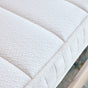 Close-up of the Eco Organic Crib Mattress by Avocado Green Mattress, featuring white, quilted organic fabric with textured detailing and visible stitching along the edge, resting on a wooden bed frame.