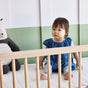 A toddler in a blue polka dot dress sits in a crib next to a stuffed animal. The crib, featuring light wooden rails and an Eco Organic Crib Mattress by Avocado Green Mattress, is set against a wall painted half white, half green.