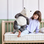 A young child sits on a bed with an Avocado Green Mattress Eco Organic Crib Mattress, smiling and holding a large plush panda toy. The bed features light-colored bedding and a wooden frame, set against a green and white wall in the background.