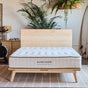 A modern bedroom featuring a wooden bed frame and an Extra Firm Mattress from Avocado Green Mattress—celebrated for its certified organic materials and heavy-duty support—alongside potted plants, a surfboard, and a partially visible bike, all set atop a patterned rug.