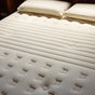 Close-up of the Avocado Green Mattress Extra Firm Mattress, a certified organic beige mattress with textured patterns and two matching pillows on top. The mattress features tufted and indented sections, highlighting its plush yet supportive extra firm design.