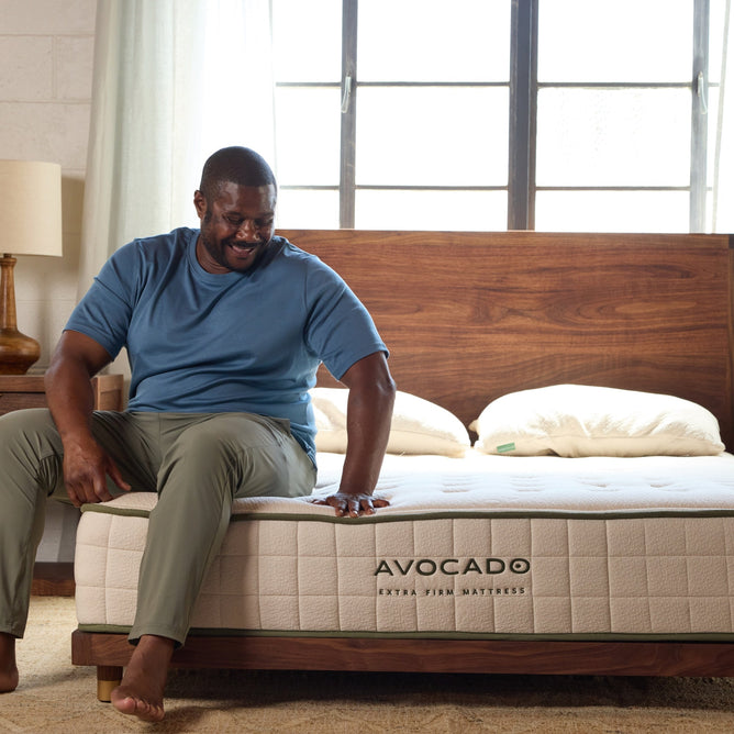 A man sits on an Avocado Green Mattress Extra Firm Mattress, pressing down on the heavy-duty certified organic mattress with one hand, in a sunlit bedroom featuring a wooden bed frame and two pillows.