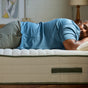 A man in a blue shirt and gray pants sleeps peacefully on his side on an Avocado Green Mattress Extra Firm Mattress in a cozy, softly lit bedroom.