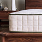 A close-up side view of the Avocado Green Mattress by Avocado Green Mattress, featuring a thick, quilted organic build with green trim on a wooden bed frame. Wooden furniture and decorative items are visible in the background.