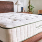 A close-up of a plush, quilted Avocado Green Mattress on a wooden bed frame in a bright, modern bedroom with a lamp, books, and a plant in the background. The certified brand name Avocado Green Mattress is visible.