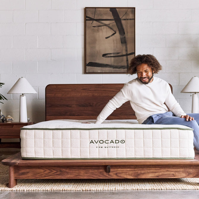 A smiling man sits on a bed with a wooden frame, touching an Avocado Green Mattress by Avocado Green Mattress. Two lamps and abstract wall art decorate the background, enhancing the serene setting as he enjoys the pressure-relieving comfort of this nontoxic mattress.