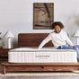 A smiling man sits on a bed with a wooden frame, touching an Avocado Green Mattress by Avocado Green Mattress. Two lamps and abstract wall art decorate the background, enhancing the serene setting as he enjoys the pressure-relieving comfort of this nontoxic mattress.