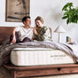 A smiling couple sits on a bed with gray sheets in a cozy bedroom. The mattress label reads Avocado Green Mattress by Avocado Green Mattress, known for its organic materials and pressure-relieving comfort. A plant and framed artwork are in the background.