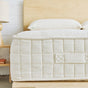 A close-up of the Luxury Organic Mattress by Avocado Green Mattress, featuring a white, quilted design with a fabric handle, placed on a wooden bed frame. In the background, a wooden nightstand with a lamp and pillows can be seen.