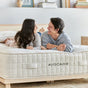A smiling couple lies on a bed, facing each other, on an Avocado Green Mattress Luxury Organic Mattress in a cozy, modern bedroom with shelves and decor in the background.