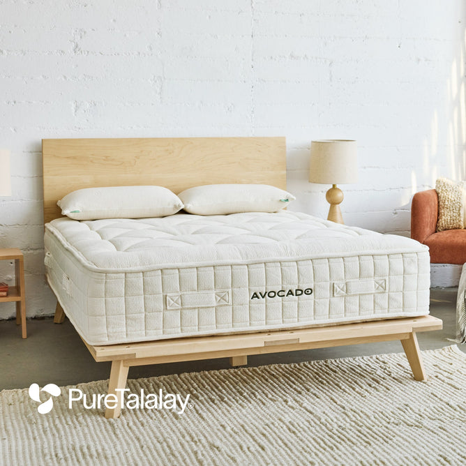 A modern bedroom with a wooden bed frame, an Avocado Green Mattress Luxury Organic Mattress, white pillows, two beige lamps, and a small orange chair. The PureTalalay logo appears in the bottom left corner.