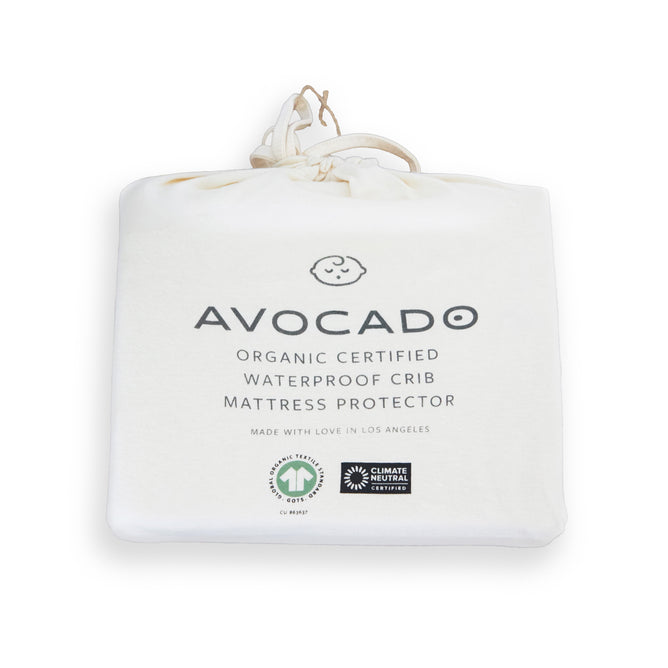 The Avocado Organic Crib Waterproof Mattress Protector comes in a white fabric bag with minimalist text and icons, drawstring closure, eco-friendly certification logos, and is MADE SAFE certified.
