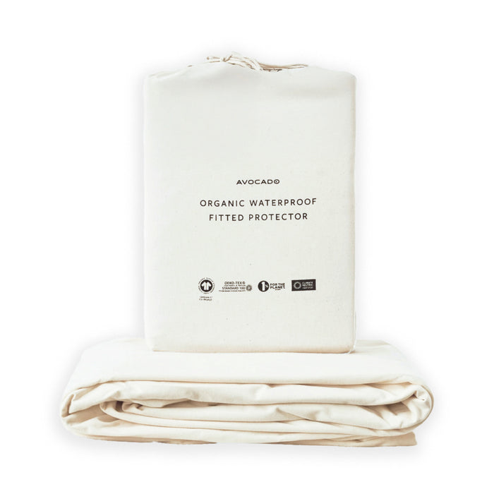 A folded cream-colored Avocado Organic Waterproof Mattress Protector is displayed with its matching storage bag, featuring minimalist black text, eco-certification logos, and a MADE SAFE Certified label on the front.