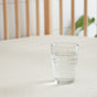 A clear glass of water rests on a light surface with a wooden chair and green plant blurred behind, evoking the freshness and purity of Avocado’s Organic Crib Waterproof Mattress Protector.