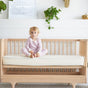 A young child sits cross-legged on a light wooden crib, topped with the Avocado Organic Crib Waterproof Mattress Protector, against a white wall adorned with green plants and framed art.