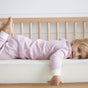 A smiling blonde-haired child in pink and white striped pajamas lies on their stomach on an Avocado Organic Crib Waterproof Mattress Protector, with a wooden crib rail behind them.