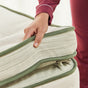 Wearing maroon pajamas, a person lifts the corner of an Avocado Green Ultra Plush Mattress by Avocado Green Mattress, highlighting its thick profile and textured white and green fabric.