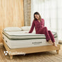 A woman in maroon pajamas sits smiling on a bed featuring the Avocado Green Ultra Plush Mattress from Avocado Green Mattress in a bright, modern bedroom. The PureTalalay logo appears in the corner.